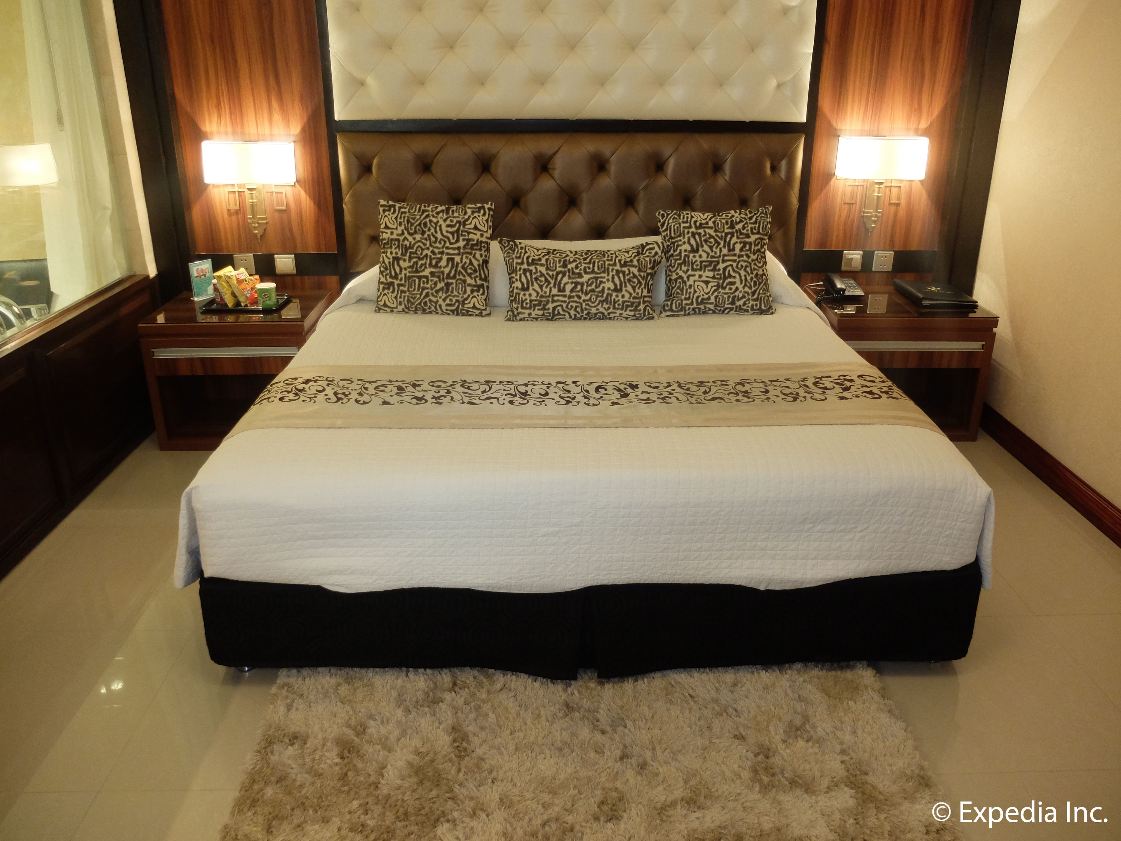 A large bed with a tufted headboard, patterned pillows, and a decorative bed runner. There are two bedside tables with lamps and a nightstand with a phone and a remote control.