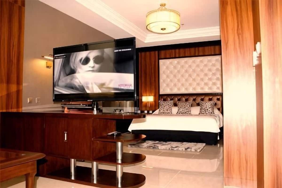 Ein Hotelzimmer mit Fernseher, einem Bett mit gestepptem Kopfteil, einer kleinen Sitzecke und einem Holzschrank.