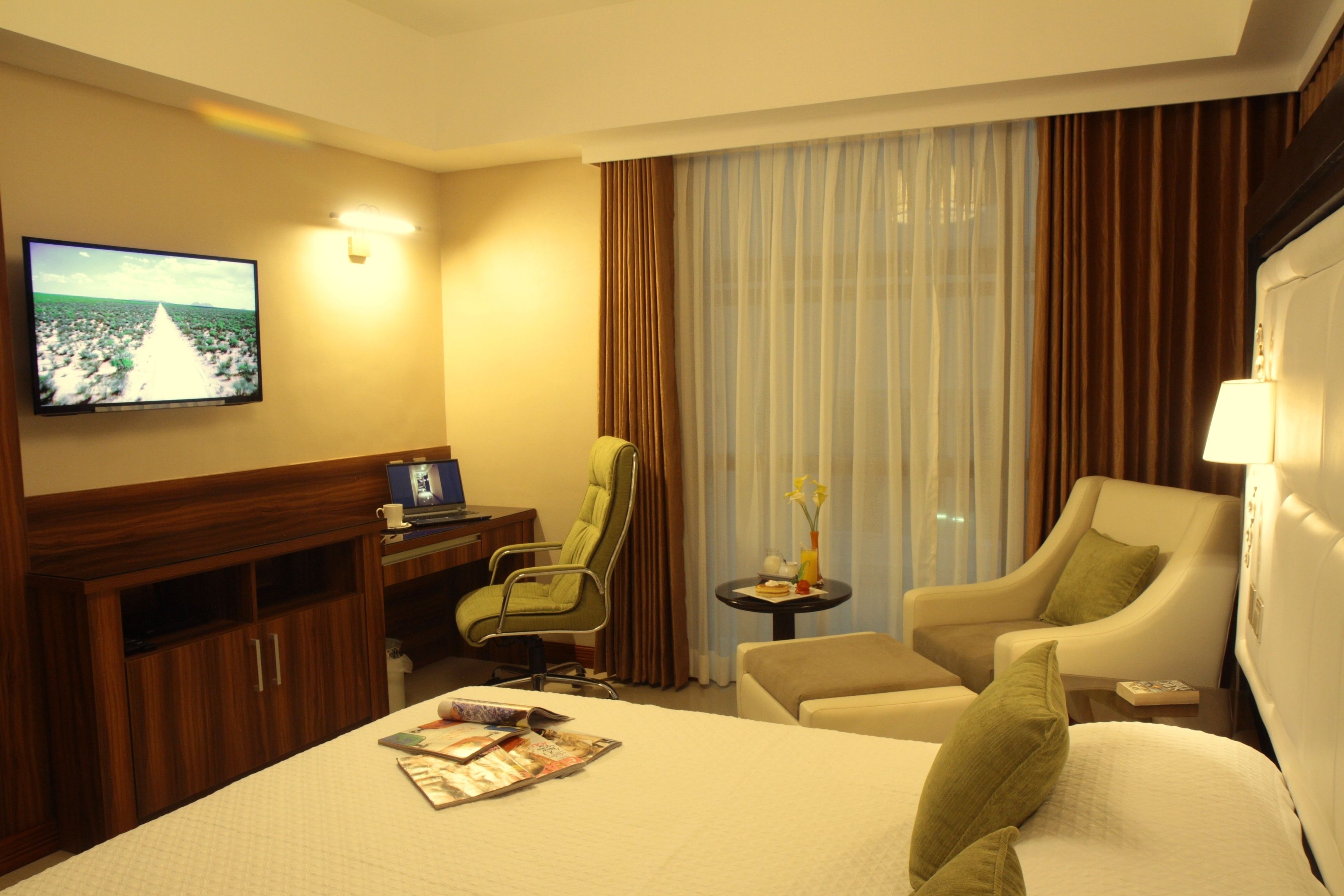 A hotel room with a large bed, a desk with a computer, a TV mounted on the wall, and a seating area with two chairs.