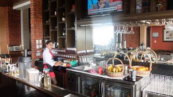A bar area with a television displaying a news program, shelves with bottles, and a bartender at the counter.