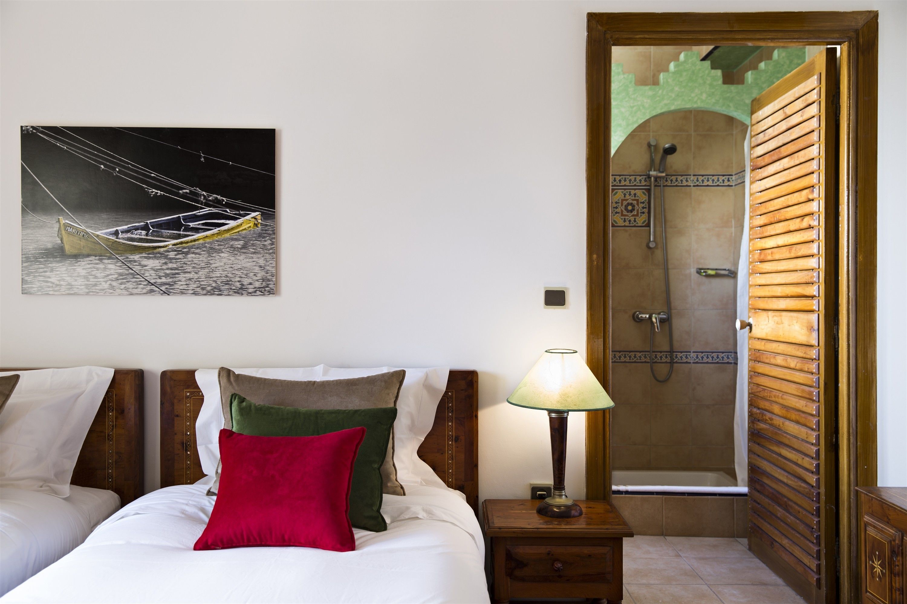 A hotel room with a bed, a nightstand with a lamp, a wooden headboard, a painting of a boat, and a shower area with a wooden door.