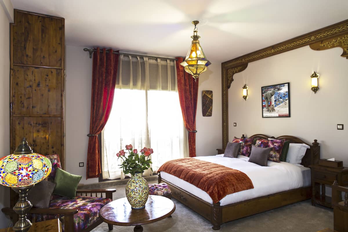 A spacious bedroom with a large bed, a wooden wardrobe, a round table with a vase of flowers, and a colorful lamp.