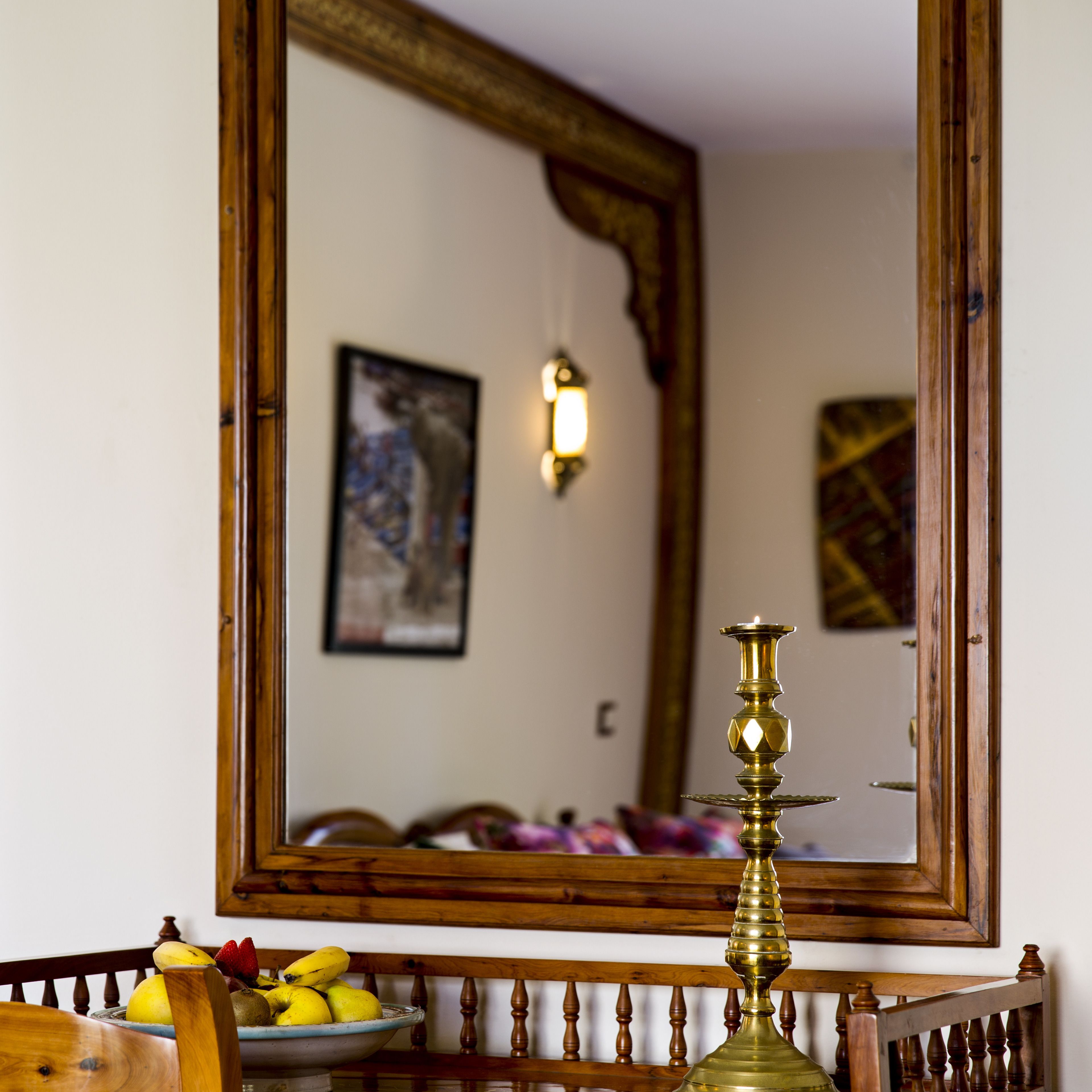 Une pièce avec un miroir à cadre en bois qui reflète un luminaire fixé au mur, un tableau encadré et une chaise en bois sur laquelle se trouve un panier de fruits.