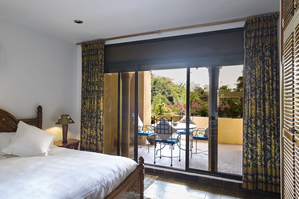 A bedroom with a large bed, a nightstand, a lamp, and a view of an outdoor patio with a table and chairs.