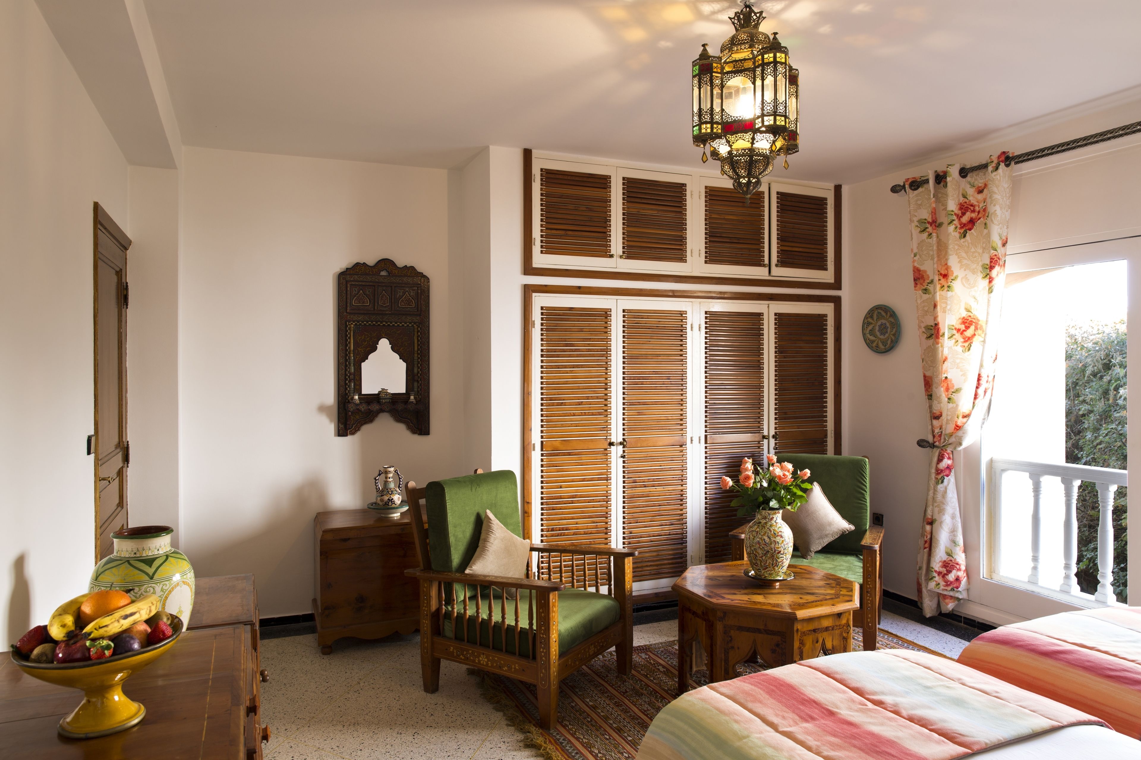 Una habitación con cama, una silla, una mesita, un jarrón con flores y una ventana con persianas.