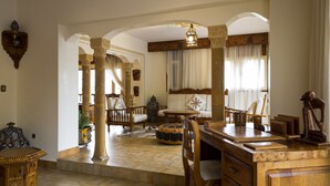 Ampla sala de estar com mesa de centro de madeira, sofá e mesa de jantar. Há colunas decorativas e um candelabro.