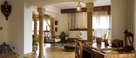 A spacious living area with a wooden coffee table, a sofa, and a dining table. There are decorative columns and a chandelier.