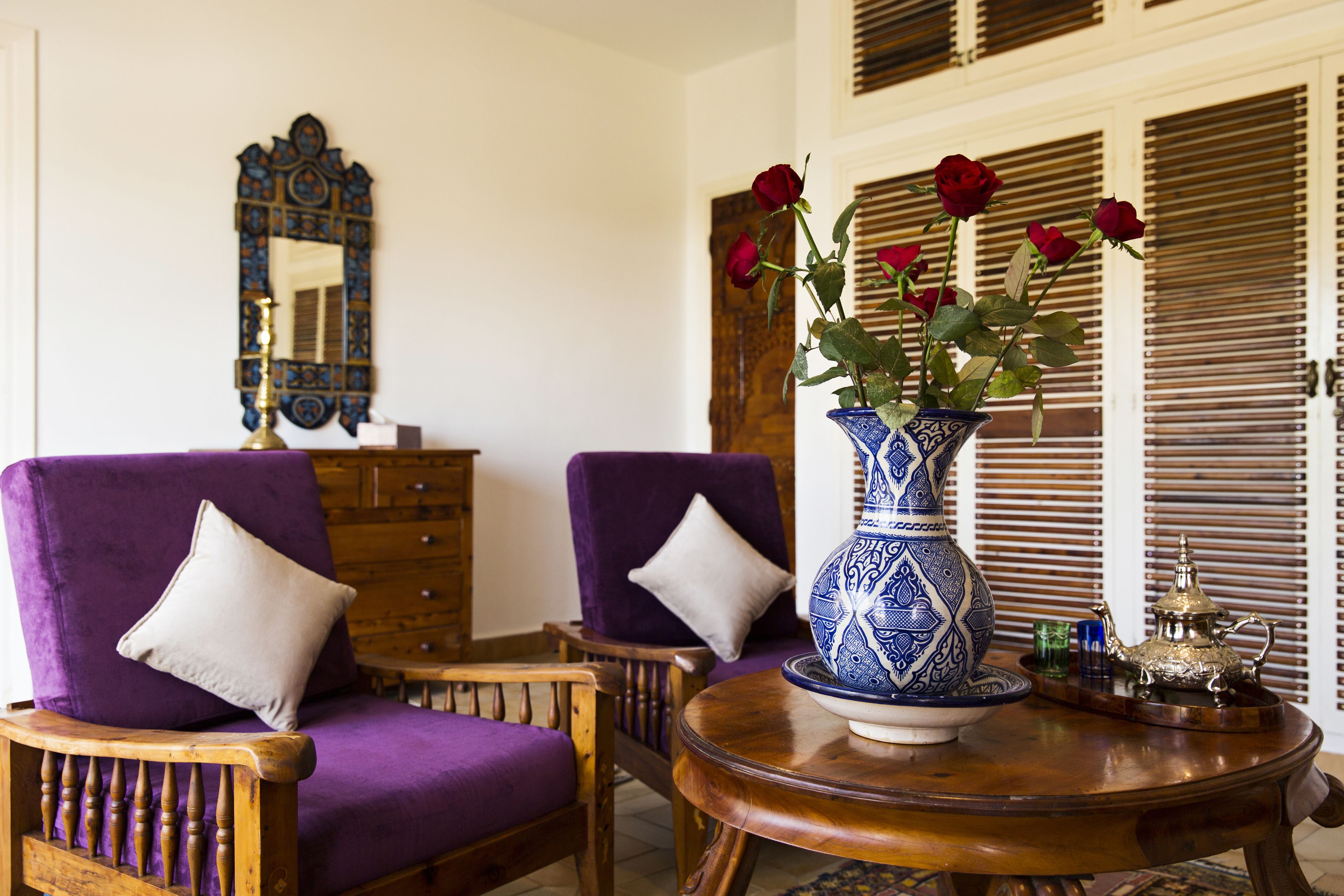 Una habitació amb dos seients de braç morats, una taula auxiliar de fusta amb un ram de roses vermelles dins d'un vas blau i blanc, i un armari de fusta.