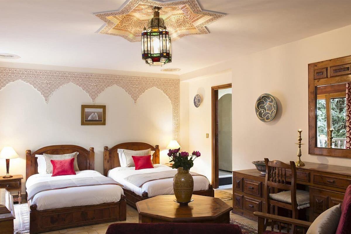 A hotel room with two beds, a wooden desk, a chair, a table, and a decorative ceiling light.