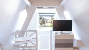 Una habitación pequeña y luminosa, con una mesa y sillas blancas, un televisor sobre un mueble blanco, y una ventana que da a palmeras y edificios.