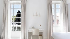 Una habitación luminosa y blanca con escritorio, silla y cama. Tres lámparas colgantes iluminan el escritorio y hay una ventana con cortinas.
