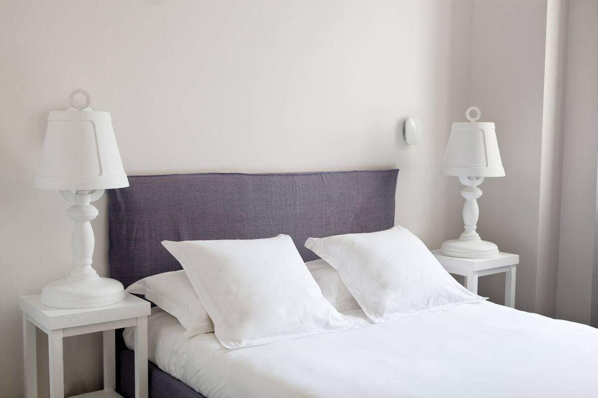 Een net opgemaakt bed met witte lakens en kussens, met aan weerszijden twee witte nachtkastjes met lampen, tegen een effen muur.