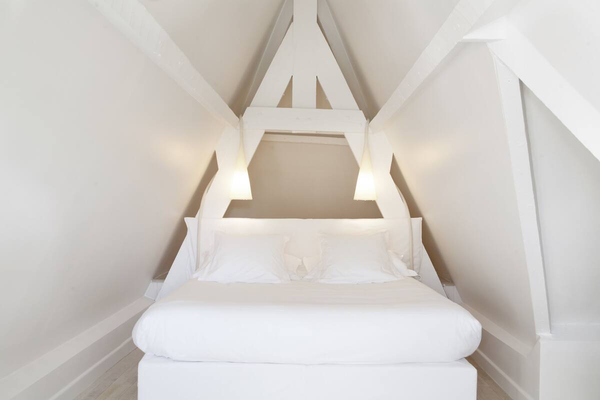 Een witte, driehoekige slaapkamer op zolder, met een bed in het midden onder een schuine hemel.