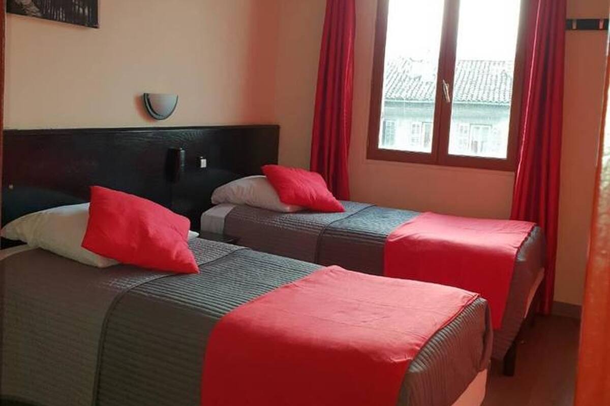 A hotel room with two beds, red curtains, a painting on the wall, and a window with a view of buildings.