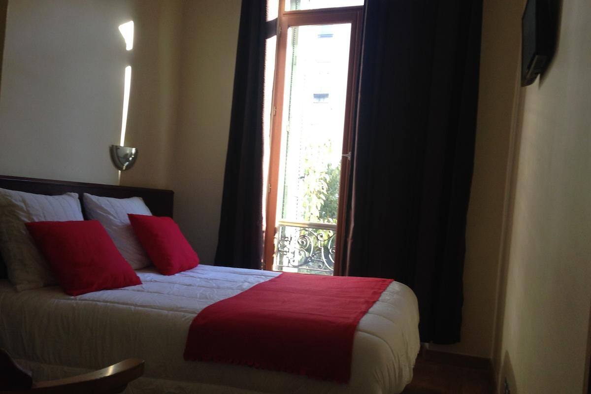 A hotel room with a bed, a nightstand, a window with curtains, and a view of the outside.