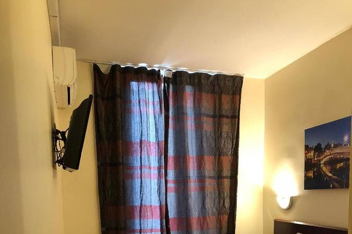 A bedroom with a bed, a headboard, a nightstand, a wall-mounted lamp, and a window with curtains.