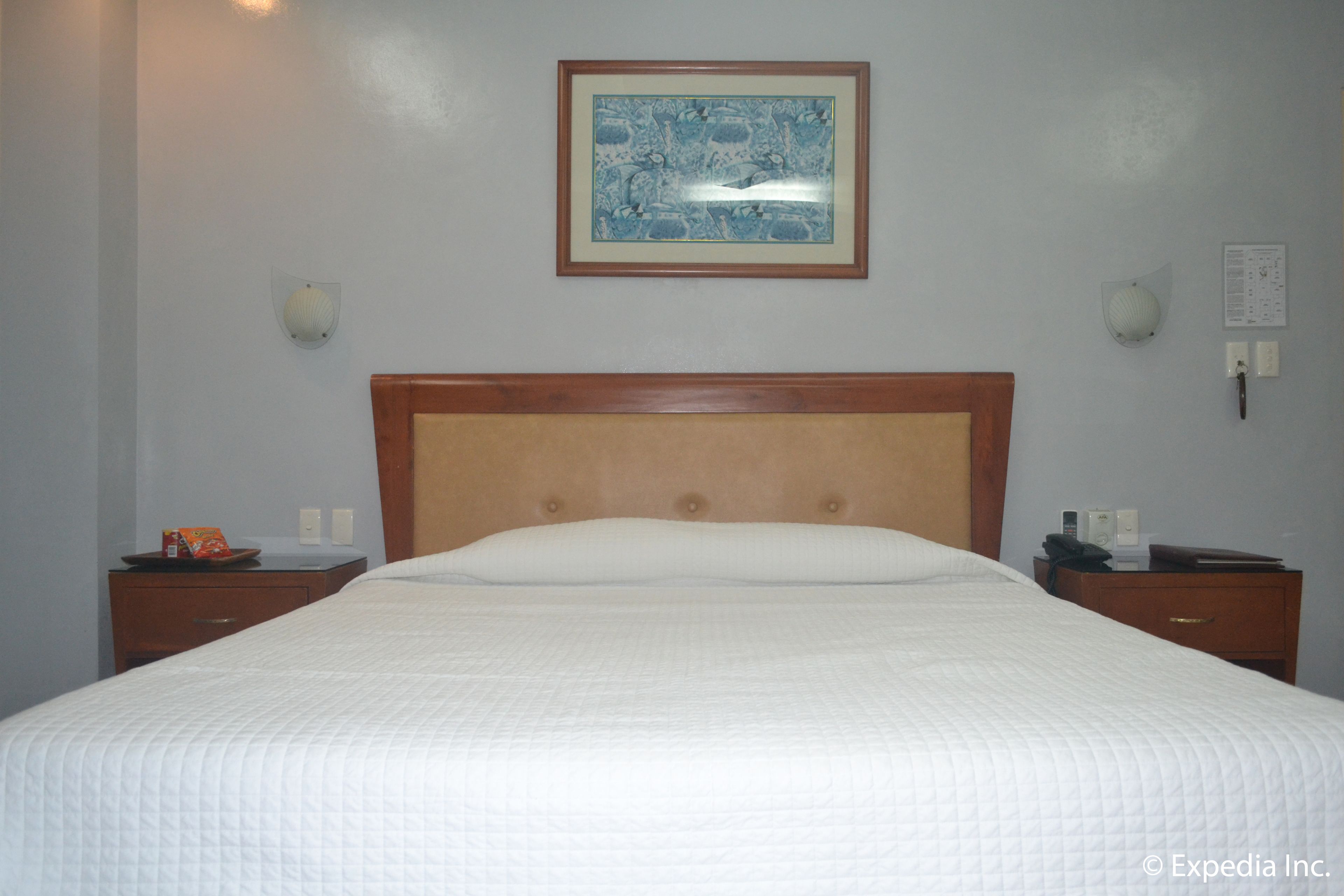 A hotel room with a large bed, wooden headboard, two bedside tables, a framed artwork on the wall, and a nightstand with a lamp.