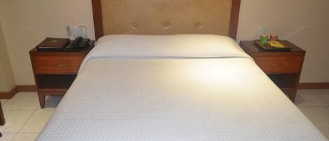 Una habitación de hotel con una cama grande, cabecera de madera, dos mesitas de noche y un atril con un teléfono y un libro.