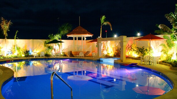 Uma área de piscina à noite, com um gazebo, espreguiçadeiras e palmeiras.