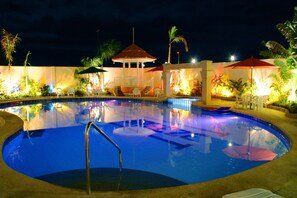 Zona de piscina iluminada con un quiosco, sillas de descanso y palmeras.