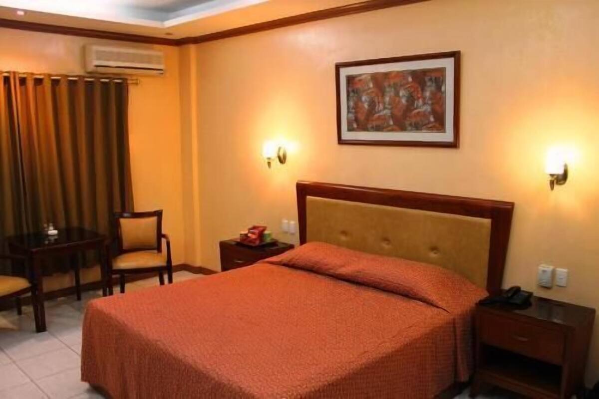 Una habitación de hotel con cama, una silla, una mesita de noche y un cuadro en la pared.