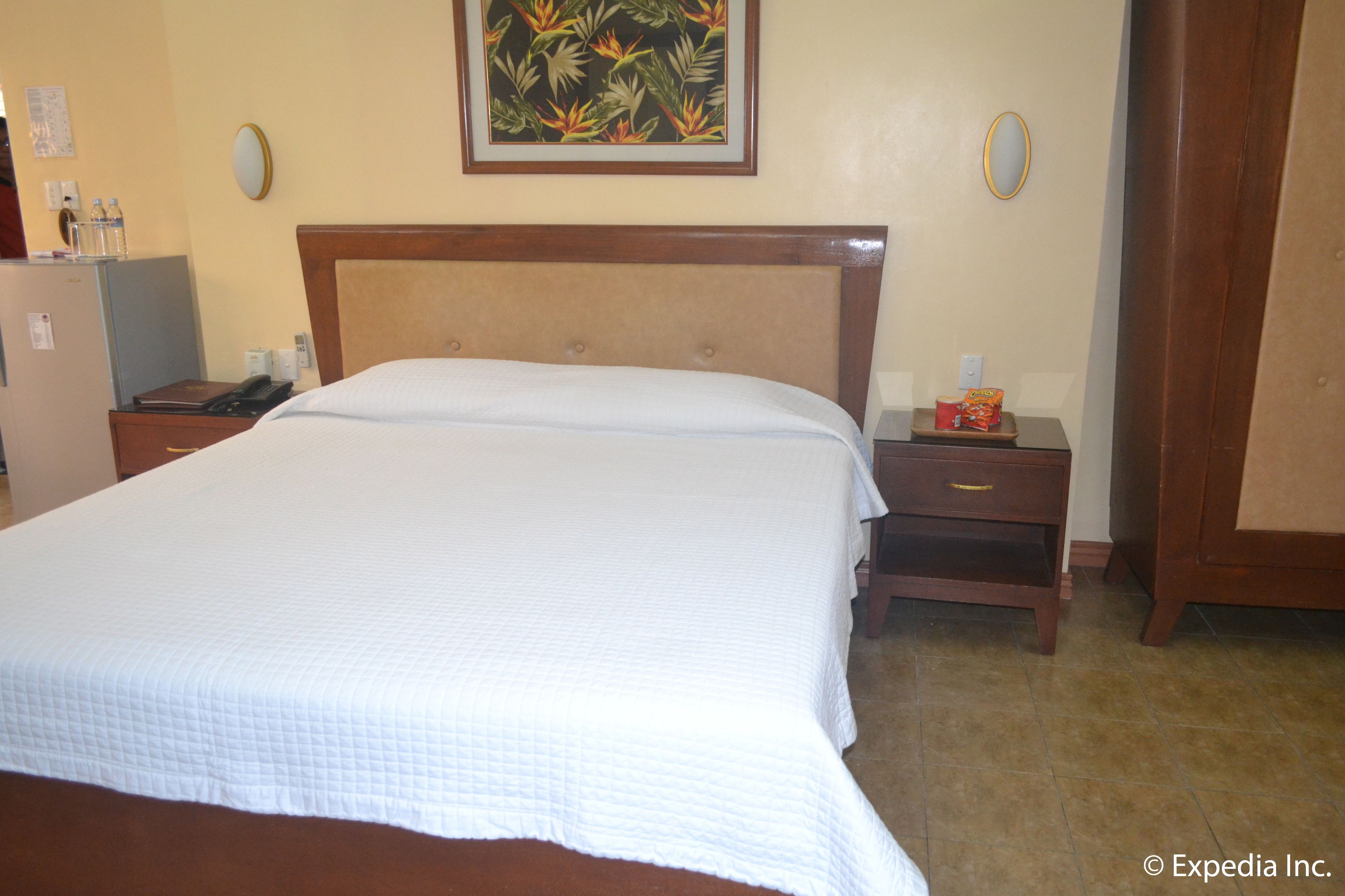 A hotel room with a large bed, a wooden headboard, a nightstand with a phone and a red box, and a painting on the wall.