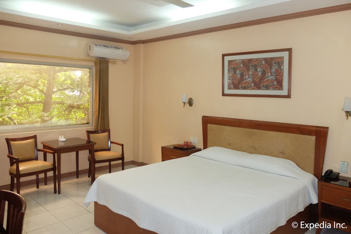 Habitación de hotel con cama, escritorio, dos sillas, ventana con cortinas, unidad de aire acondicionado, ventilador de techo e imagen en el marco en la pared.
