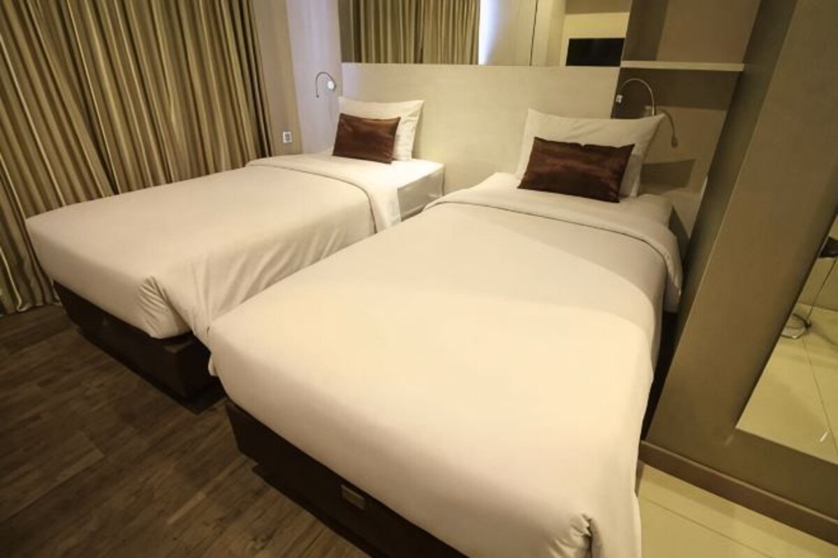 A hotel room with two beds, a mirror, a wooden floor, and a window with curtains.