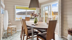 Un comedor con una mesa redonda, seis sillas, un florero con flores y vistas a un lago y montañas a través de amplios ventanales.