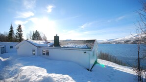Una cabaña blanca con chimenea, cerca de un lago con montañas nevadas al fondo.