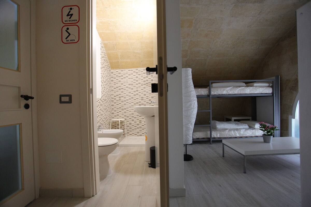 A modern bathroom with a toilet, sink, and shower. There is a bunk bed with a mattress and pillows. A potted plant is on a table.
