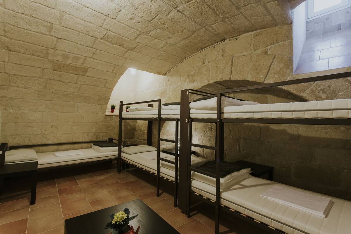 A dormitory room with bunk beds, a small table with a vase, and a window with a view of the outside.