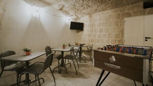 Quarto com mesa de futebol, várias mesas com cadeiras e uma televisão na parede.