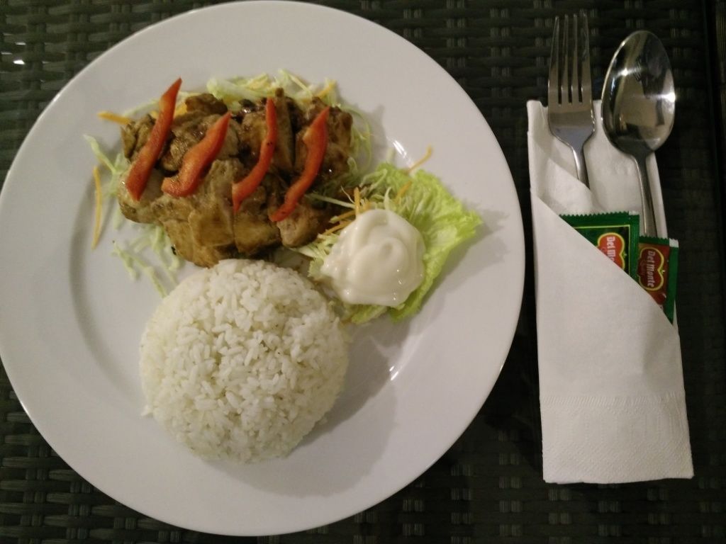 A plate with white rice, a meat dish with red peppers, and a dollop of sauce, accompanied by a napkin and utensils on a table.