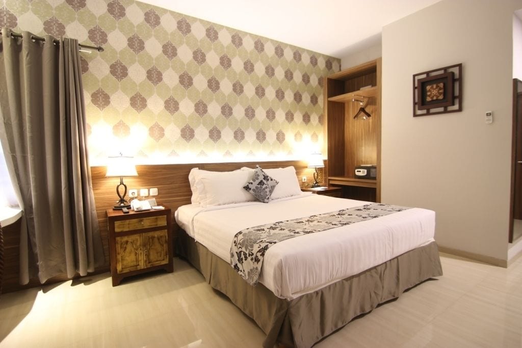 A neatly made bed with a wooden headboard, a bedside table with a lamp, and a patterned wall.