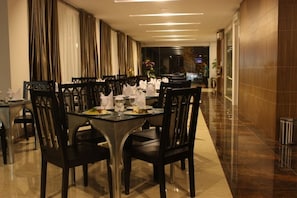 A dining area with a long table set for a meal, surrounded by chairs, with a modern interior design and a view of the street outside.