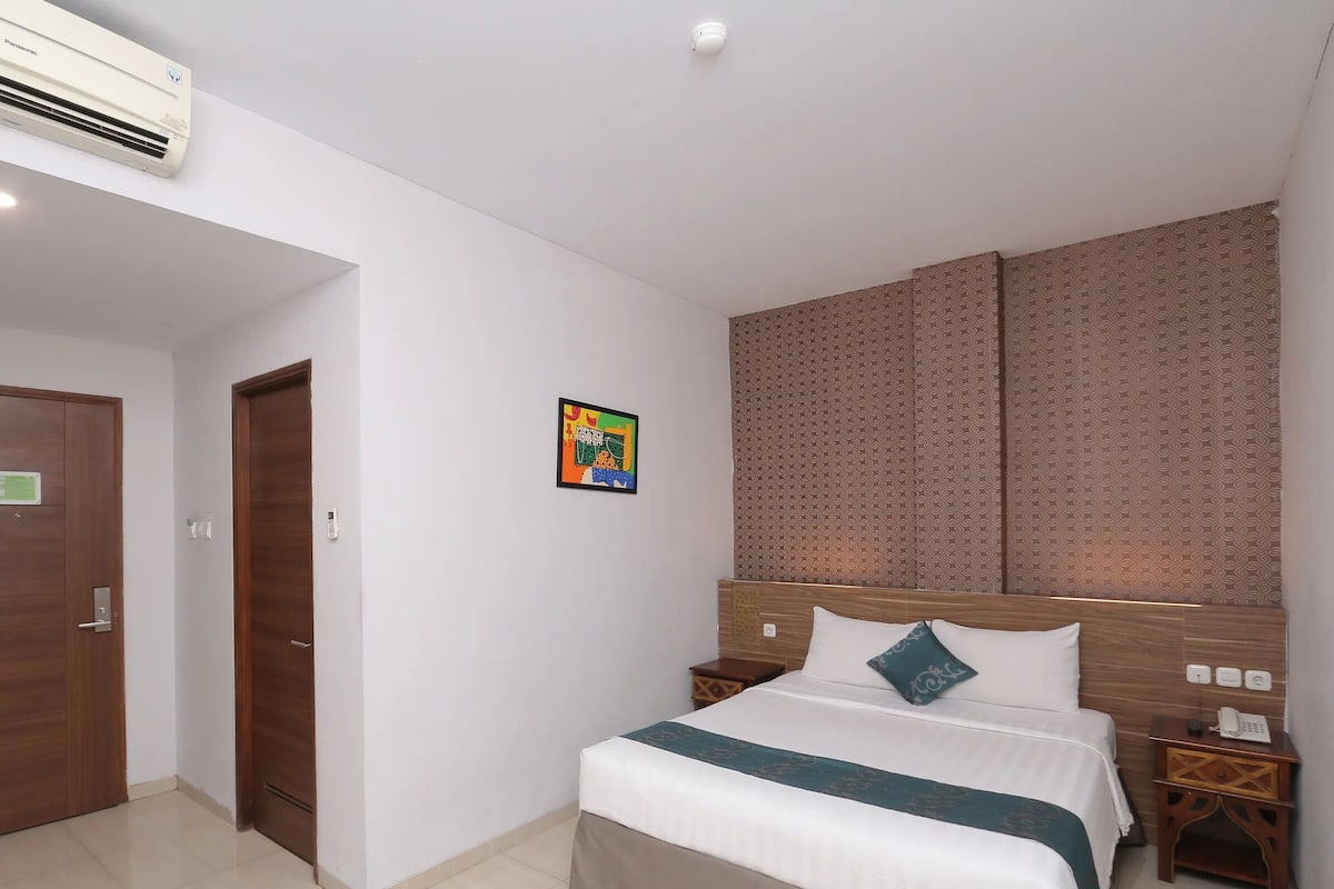 A hotel room with a bed, bedside tables, an air conditioner, a wall-mounted telephone, and a door.