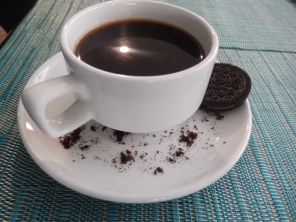 A white cup of coffee on a white saucer with a cookie and crumbs on it.