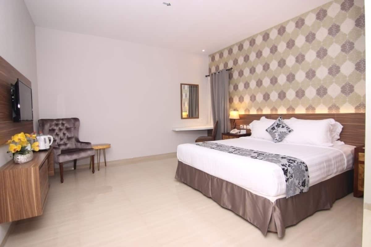 A hotel room with a large bed, a wooden nightstand, a chair, a small table, and a wall-mounted TV.