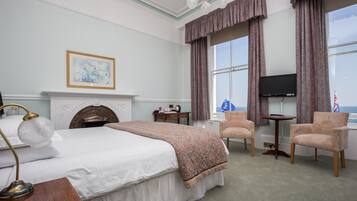 A hotel room with a bed, a desk, two armchairs, a television, and a fireplace.