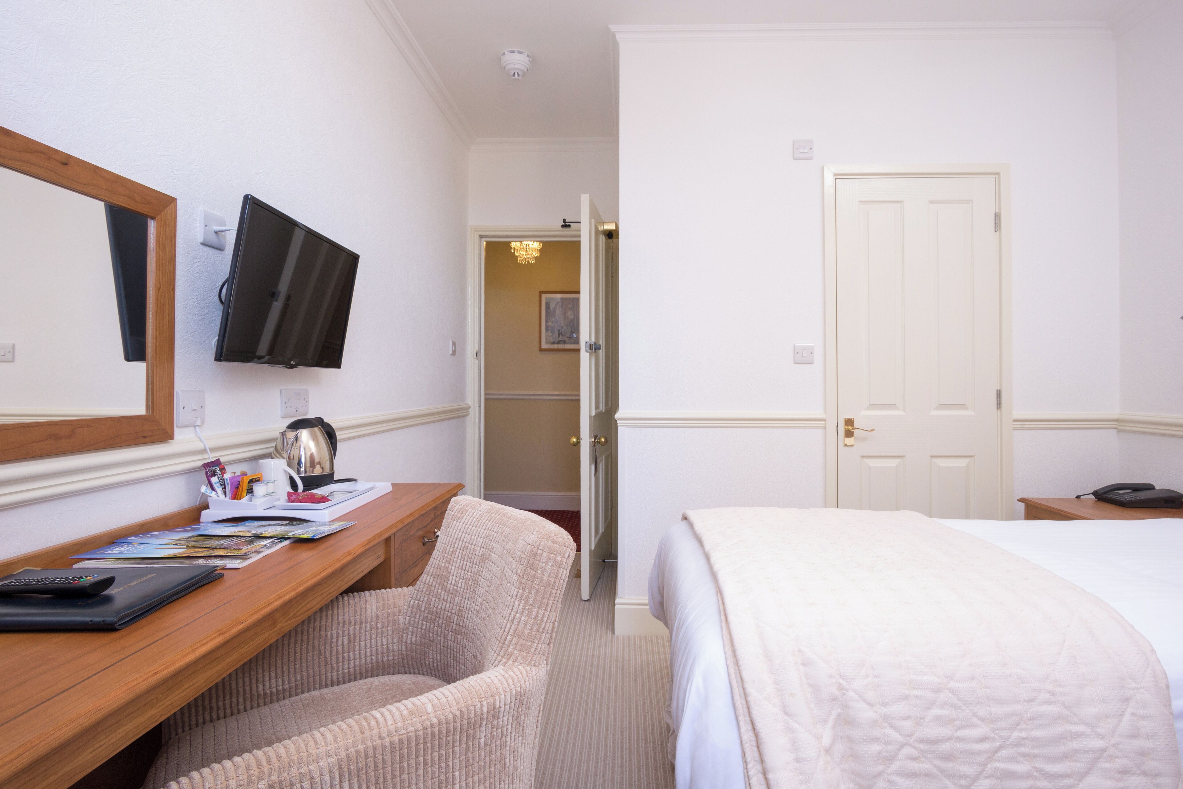 Habitación de hotel con escritorio de madera, una silla, una cama, televisor montado en la pared y una puerta que da a otra habitación.