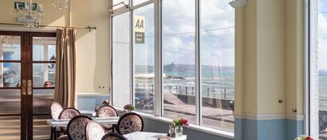 Habitación de hotel con amplios ventanales que ofrecen vista al mar y a la playa. Varias mesas y sillas dispuestas para comer.