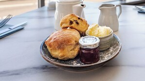 A table with pastries and condiments, a vase of flowers, and a view of the sea through large windows. - The Queens Hotel (Penzance)