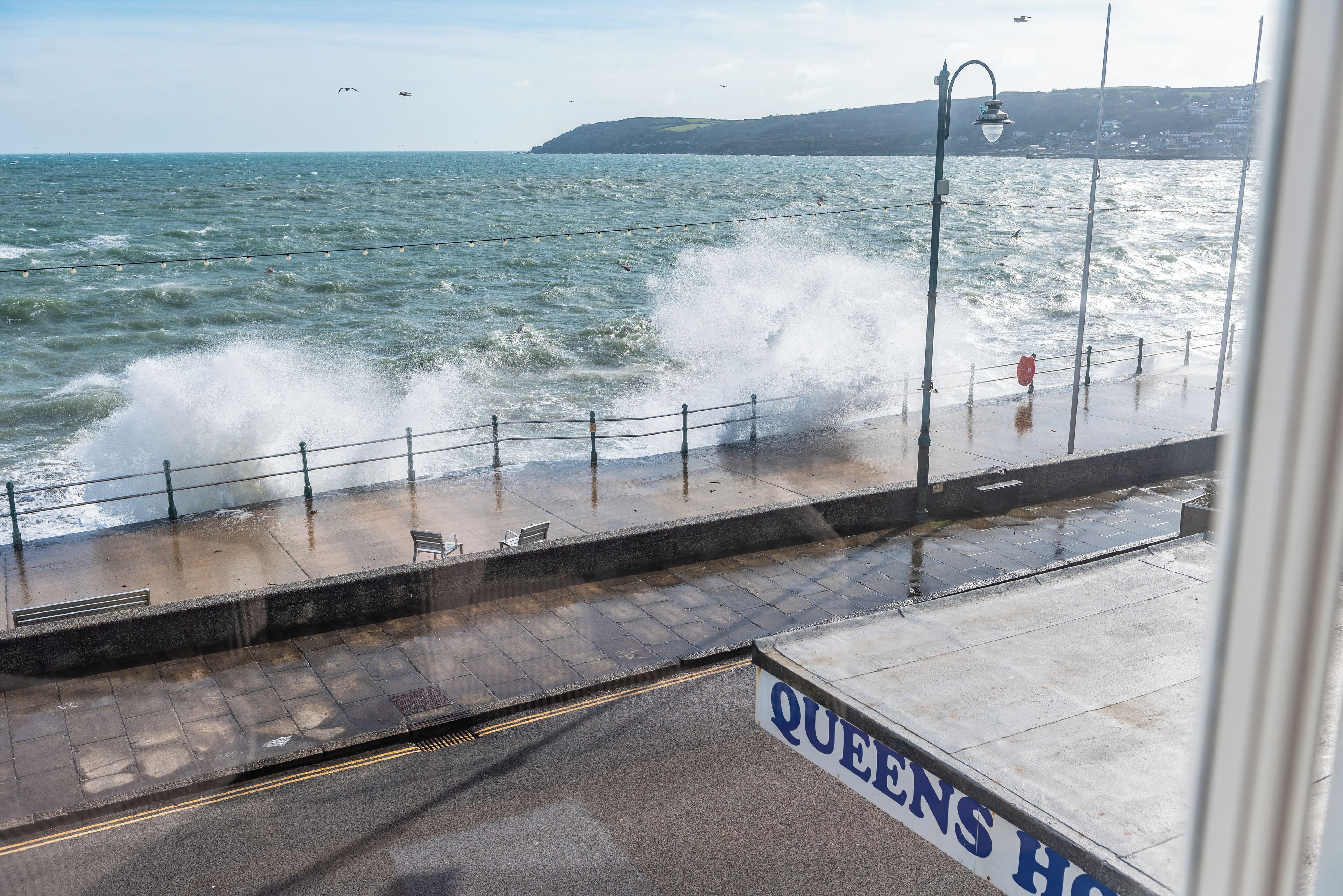 A coastal walkway with a railing, a sign reading "QUEENS HOSPITAL," and waves crashing onto the shore.