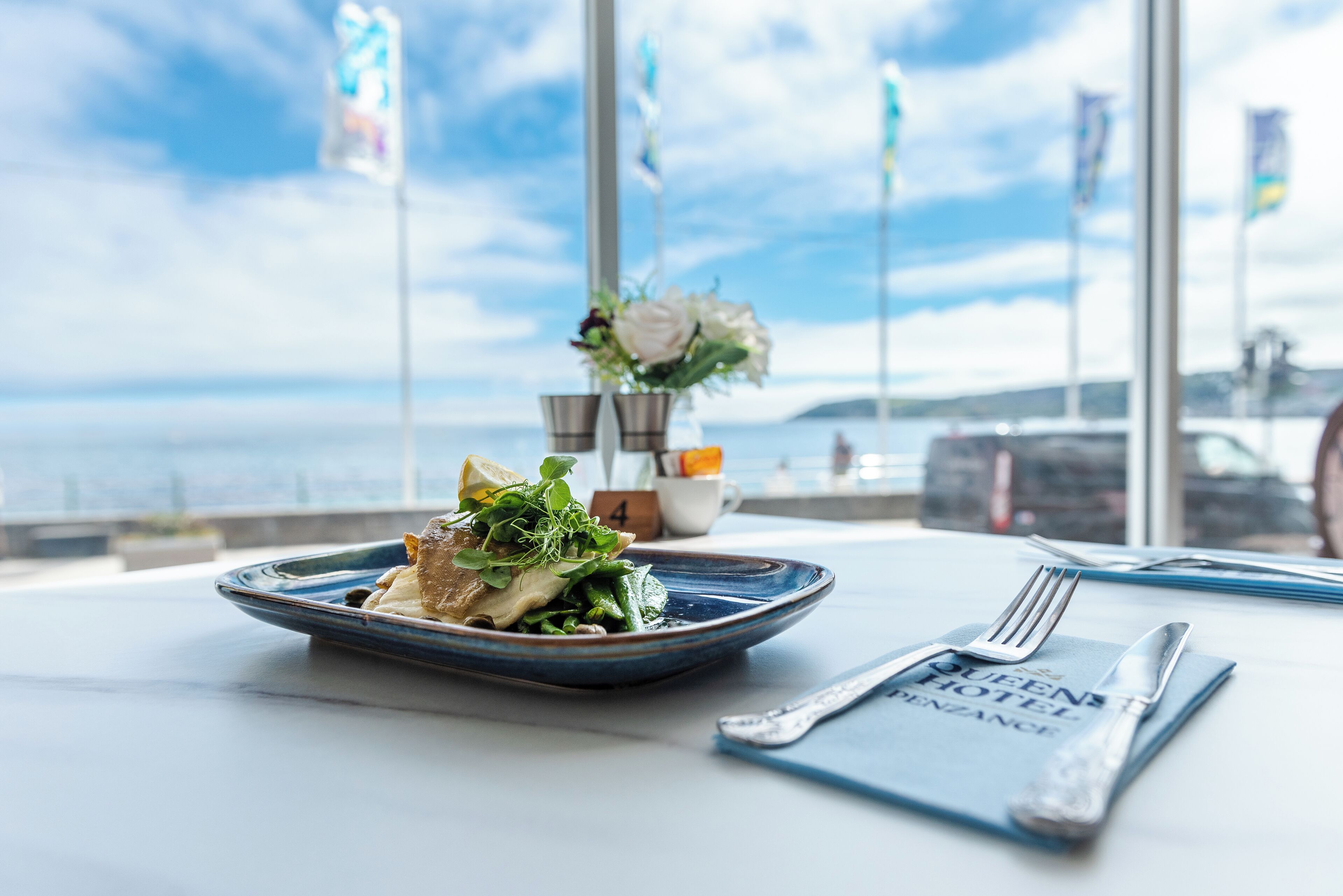 A table with a plate of food, a fork, a knife, and a menu. There is a window with a view of the sea and flags in the background.