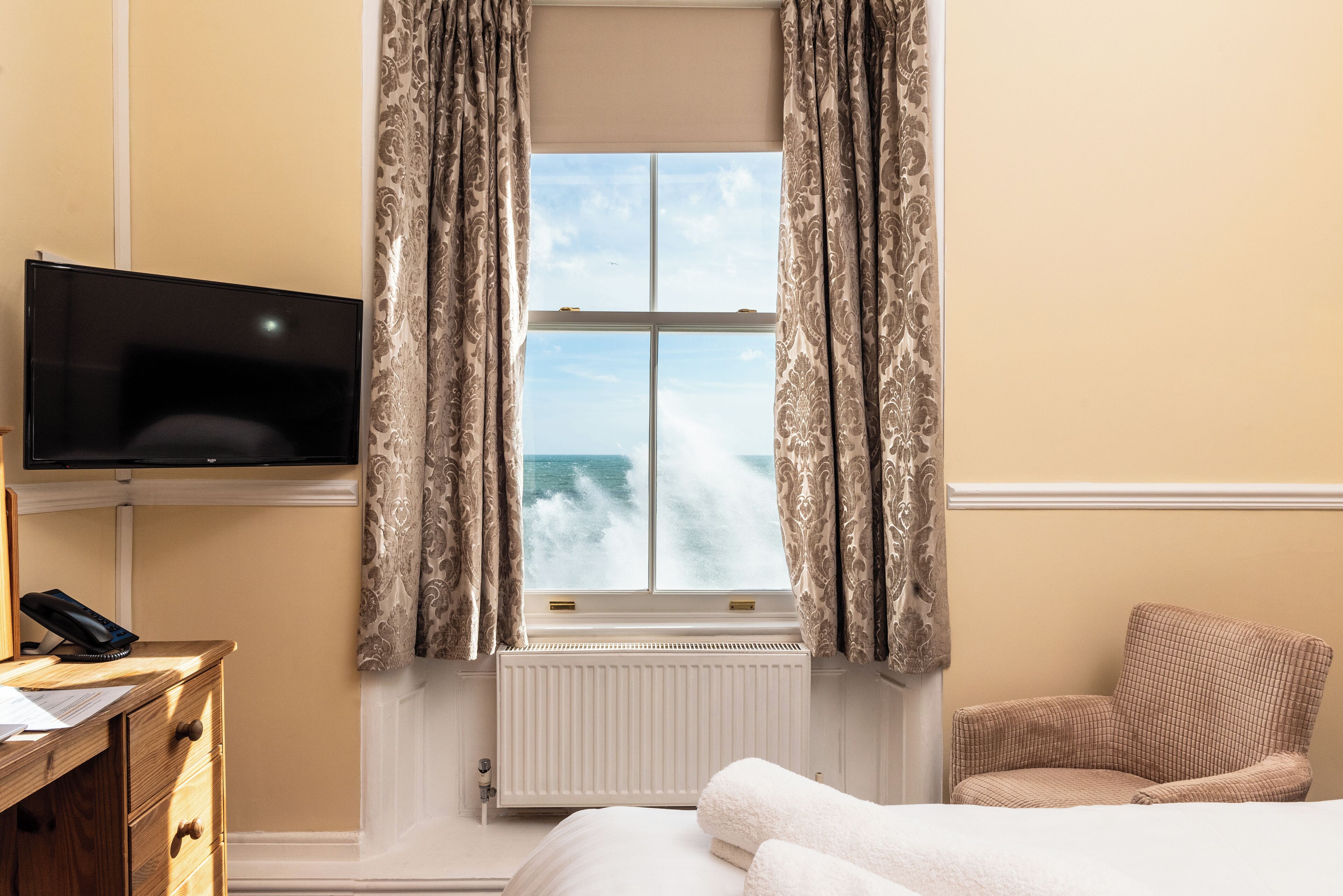 Habitación de hotel con cama, una silla, un escritorio con teléfono, televisión y ventana con cortinas.