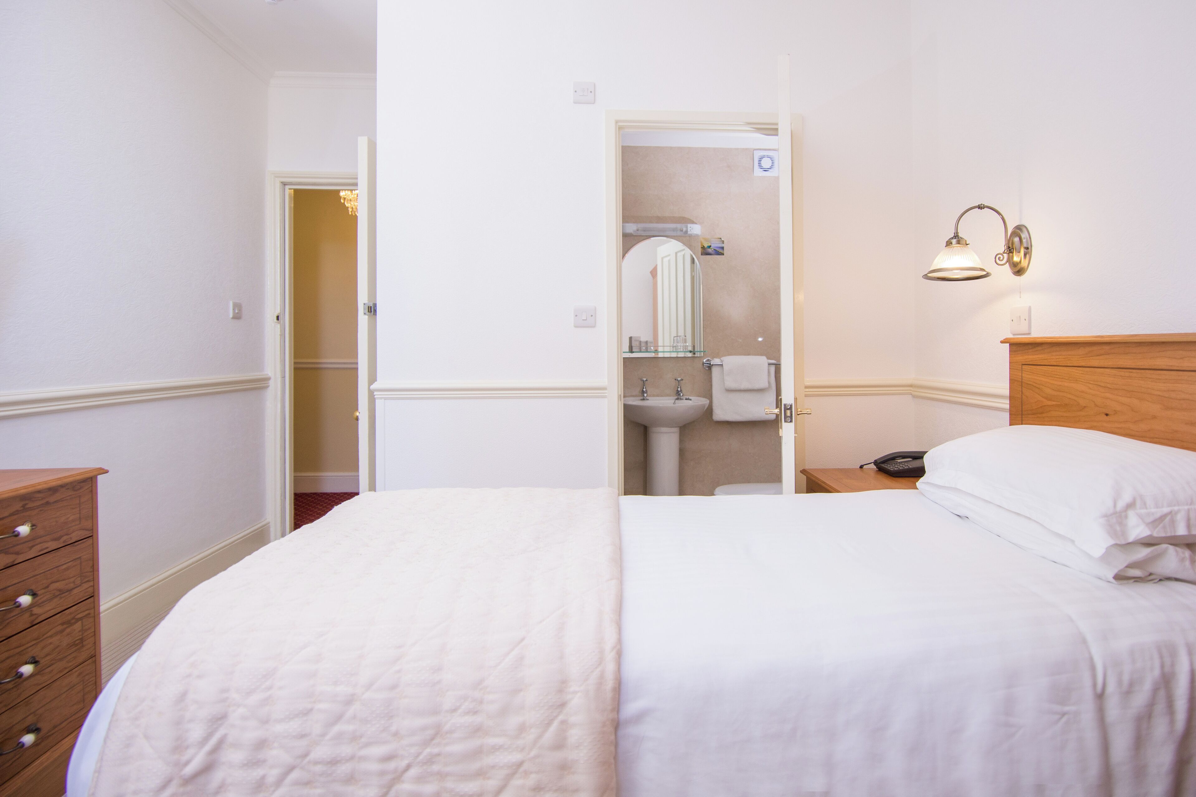 Un dormitorio con cama, cómoda de madera, mesita de noche con lámpara, y una puerta abierta que da a un baño con lavabo y espejo.