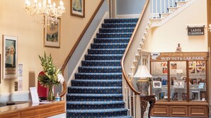 A grand staircase with a blue carpet, a reception desk with a vase of flowers, and a sign for the ladies' cloakroom. - The Queens Hotel (Penzance)