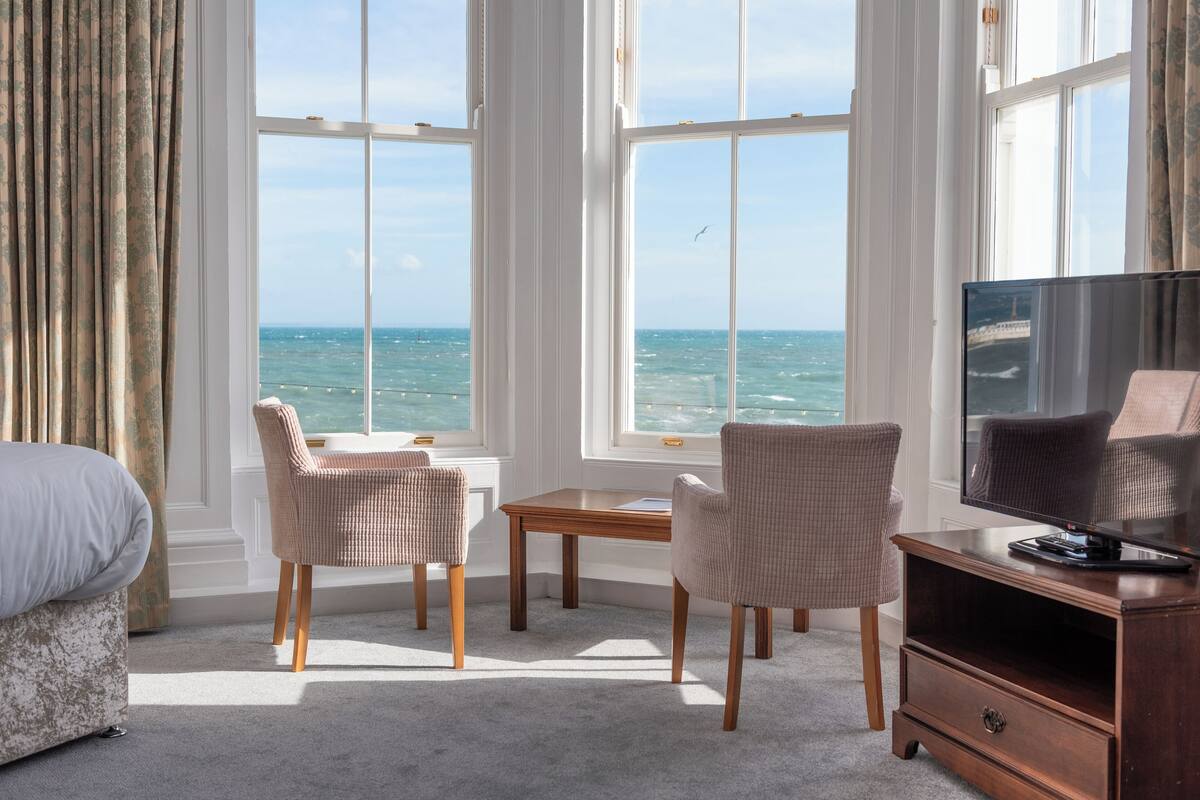 Una habitación con cama, dos sillas, una mesita, televisor sobre un soporte de madera y ventanas amplias con vista al mar.