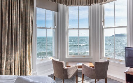 A room with a large window overlooking the sea, two chairs, a small table, and a bed.